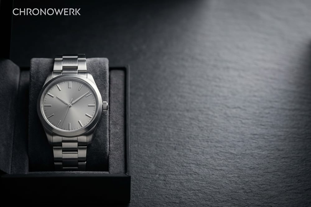 Edelstahl-Armbanduhr mit schriftfreiem silbergrauem Zifferblatt im halb geöffneten schwarzen Etui, rechts weich beleuchteter dunkler Schiefer